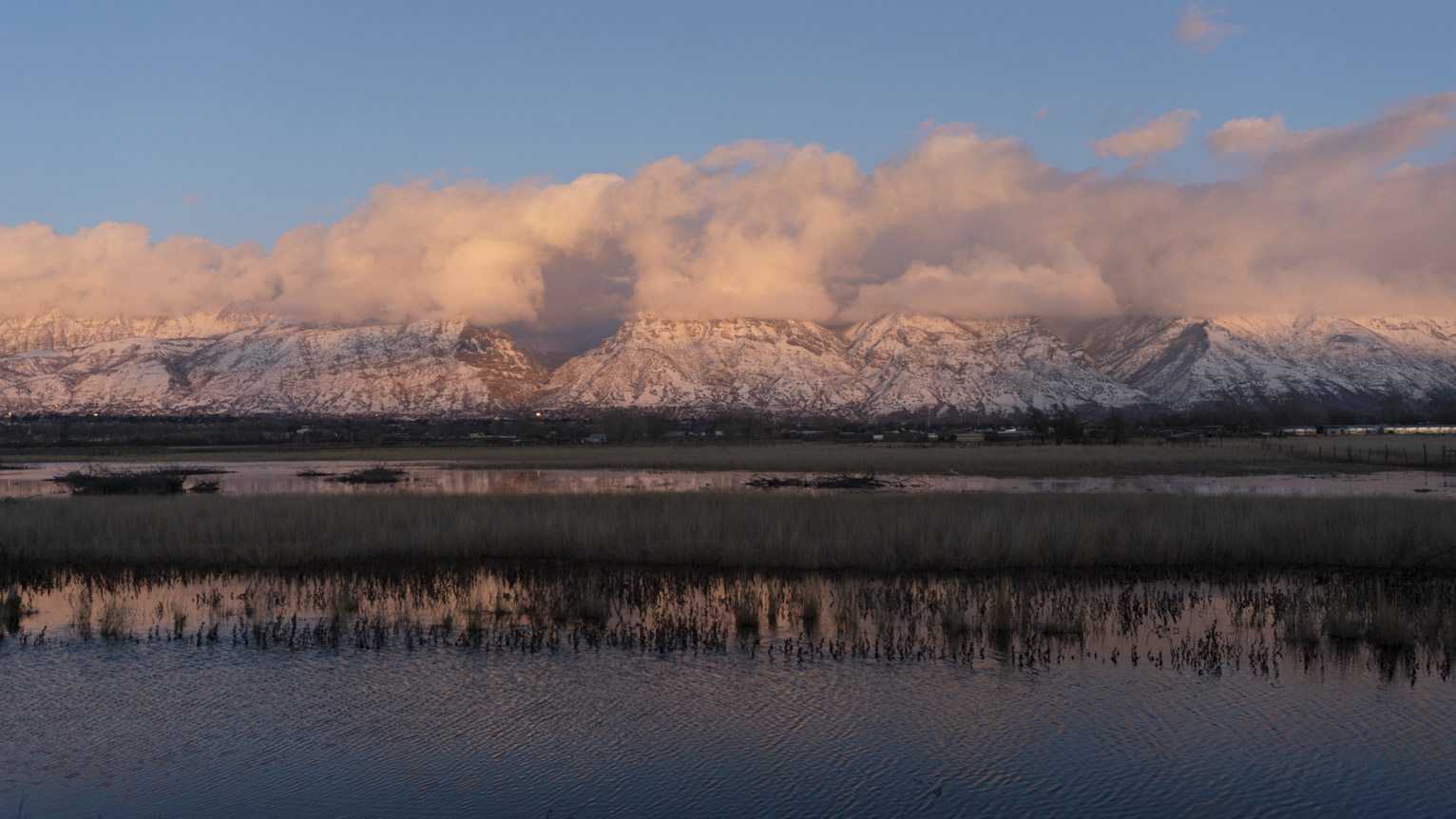 Blue sky above pinky clouds above pinky snowy mountains above their reflection in wind wrinkled water